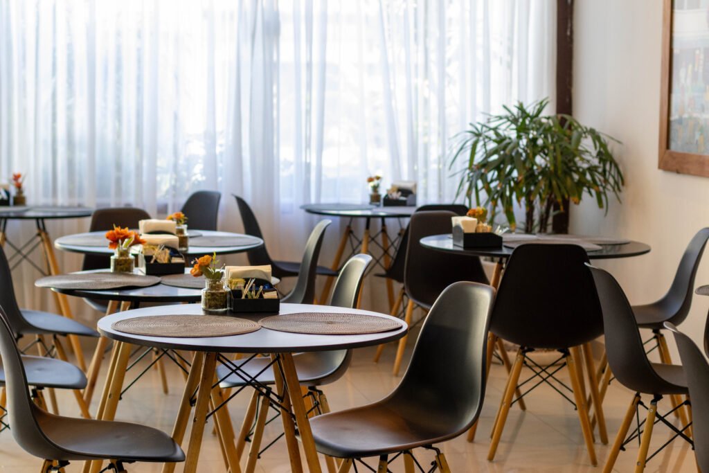 Salão de restaurante de hotel iluminado naturalmente, com mesas redondas, cadeiras modernas e arranjos de flores. Fotografia de arquitetura comercial.