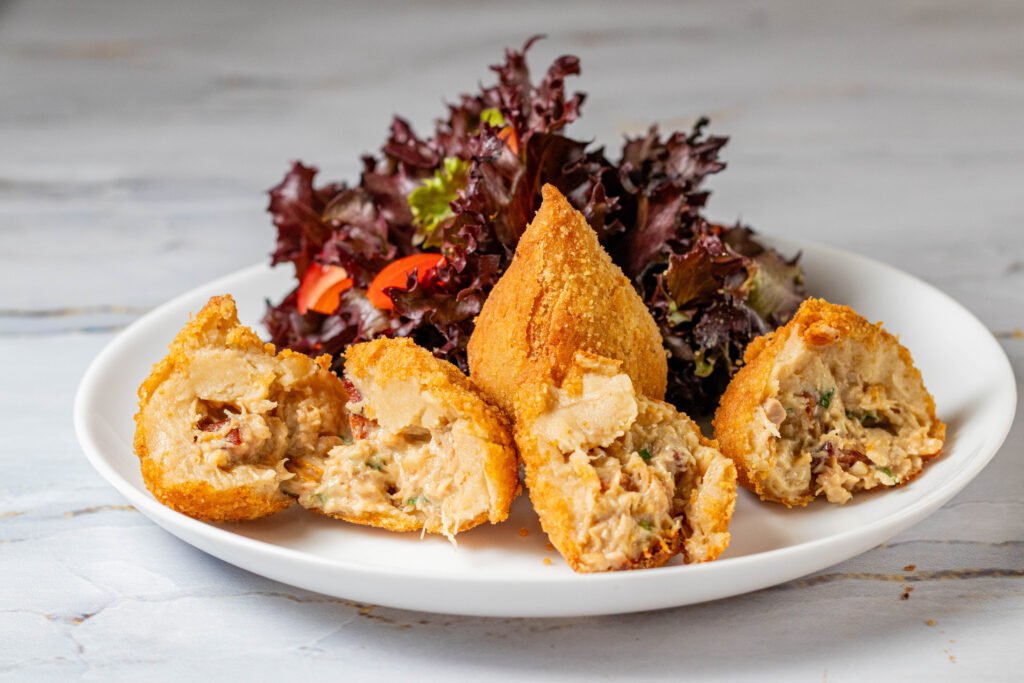 Porção de coxinhas fritas cortadas ao meio mostrando recheio cremoso e suculento, servidas com salada ao fundo. Fotografia de alimentos para marketing digital em Salvador e região metropolitana.