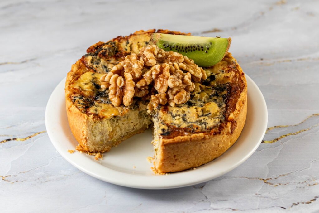 Quiche salgada inteira com uma fatia cortada, decorada com nozes e kiwi sobre prato branco. Produção de fotos para cardápios e marketing de confeitarias e restaurantes na Bahia.